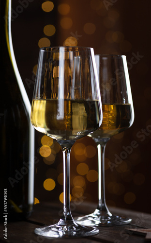 Two glasses of white wine and a bottle on a wooden table and lights garlands behind