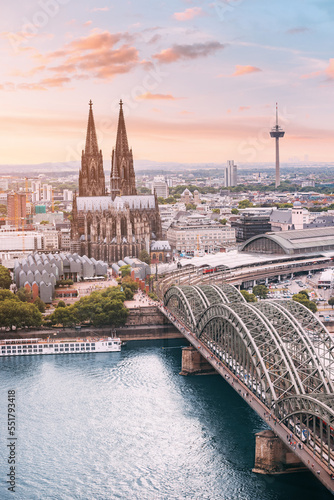 Картината върху платно Cologne Aerial vertical view with trains move on a bridge over the Rhine River on which cargo barges and passenger ships ply