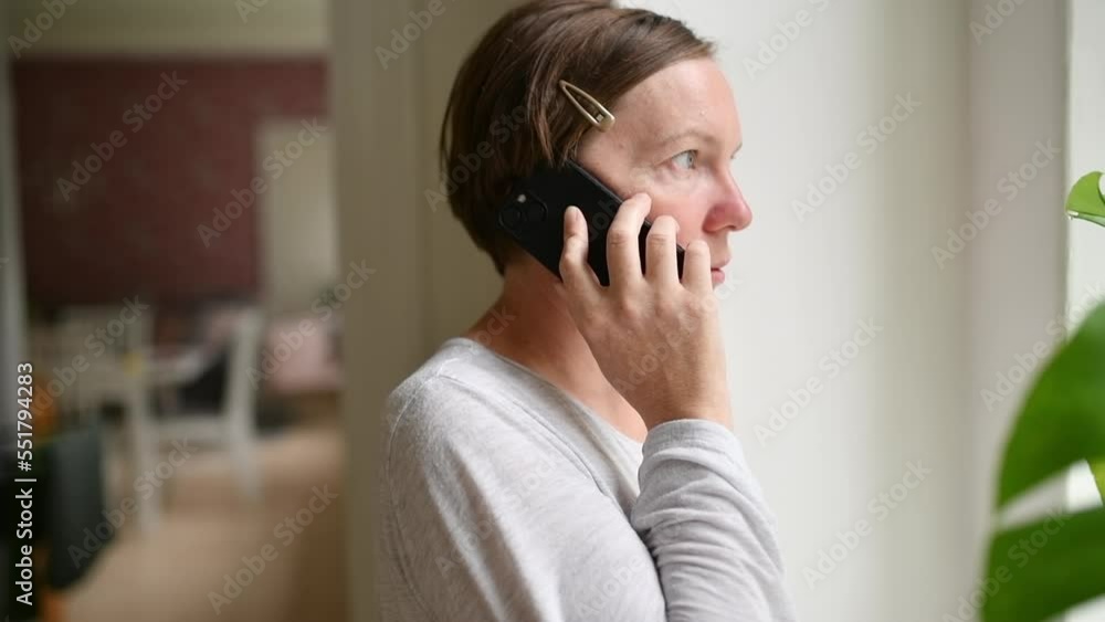 Sad alone woman talking on mobile phone while looking out the window of living room. Casual mid-adult brunette with serious facial expression. Sad alone woman talking on mobile phone while looking out the window of living room. Casual mid-adult brunette with serious facial expression.