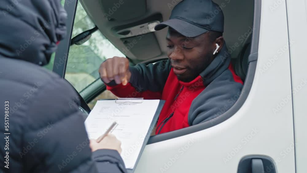 Young adult black delivery guy sitting in driver seat of white van leaning out of window to check papers signed by client. High quality 4k footage Young adult black delivery guy sitting in driver seat of white van leaning out of window to check papers signed by client. High quality 4k footage