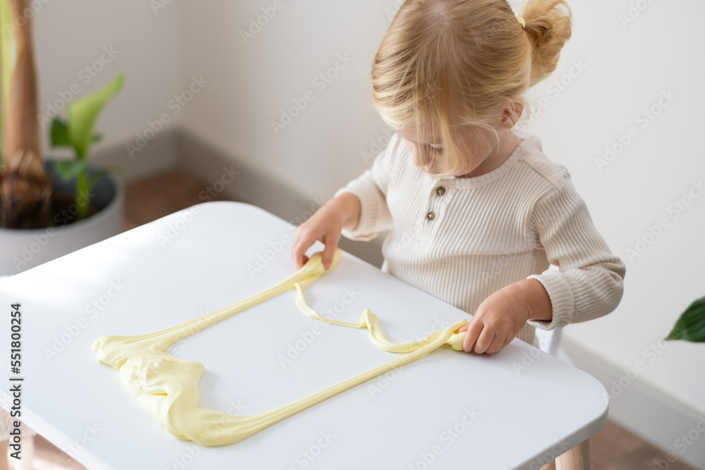 Cute blonde serious girl,toddler playing with yellow sticky slime indoors at home on white background.Early kid development, messy game, fun experiment concept.Copy space.