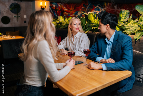 Handsome bearded male in suit and two pretty blonde women sitting restaurant talking smiling making toasts having fun, celebrating birthday. Group of happy friends drinking and toasting wine in bar.