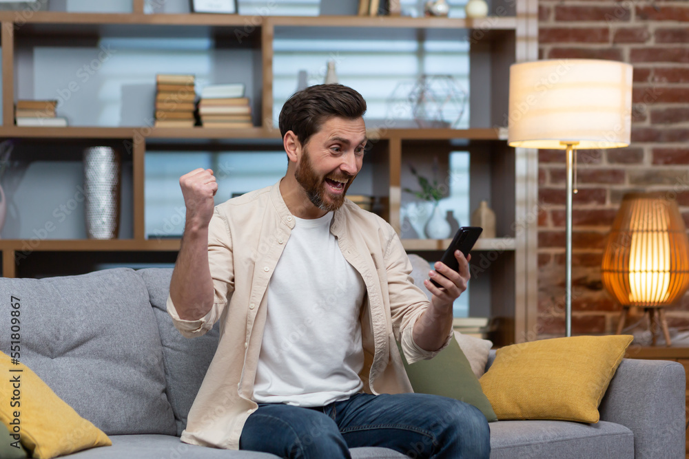 Man at home on sofa using smartphone, celebrating victory in living room reading happy news notifications from phone online holding hand up gesture of triumph and success sitting on sofa.