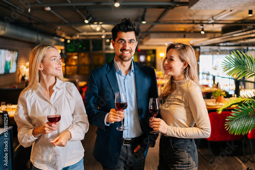 Portrait of cheerful man and two blonde women friends holding glasses of red wine, looking smiling at each other, standing posing in restaurant. Happy young male and female enjoying nice dinner.