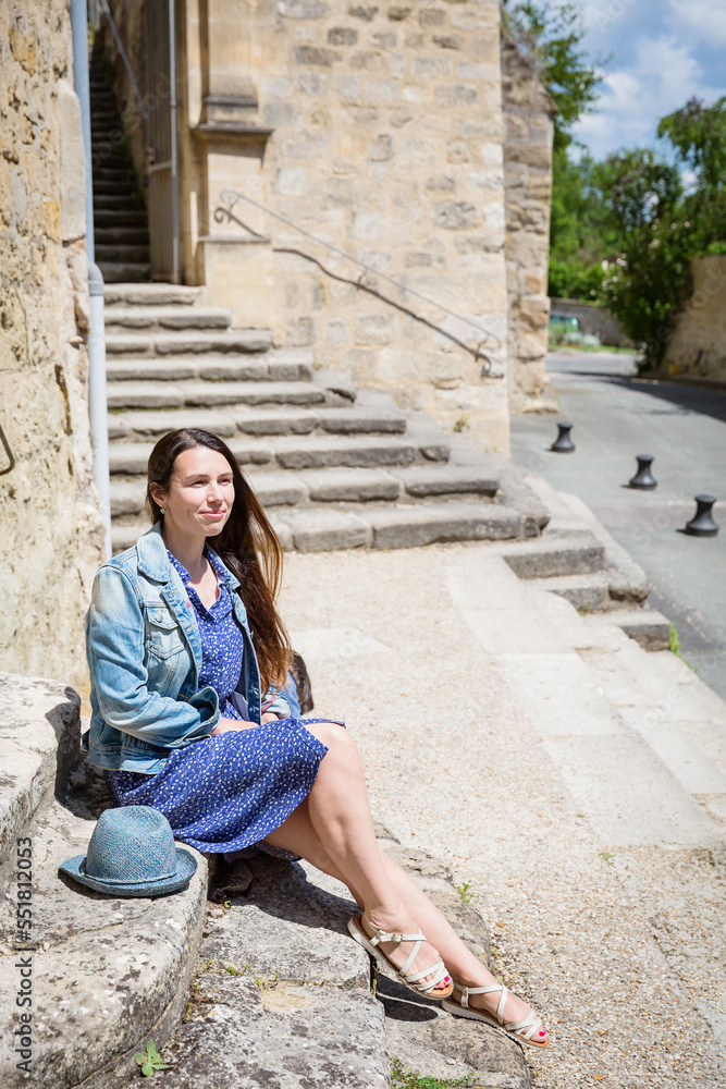 Attractive young woman sitting and posing with blue straw hat and big shopping bag on ancient stone stairs outdoor in old french city