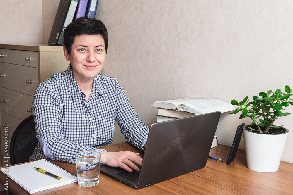 Middle-aged brunette woman sits at a desk in front of a laptop. A woman with a short haircut works with a laptop and textbooks. The woman looks into the camera. Middle-aged brunette woman sits at a desk in front of a laptop. A woman with a short haircut works with a laptop and textbooks. The woman looks into the camera.