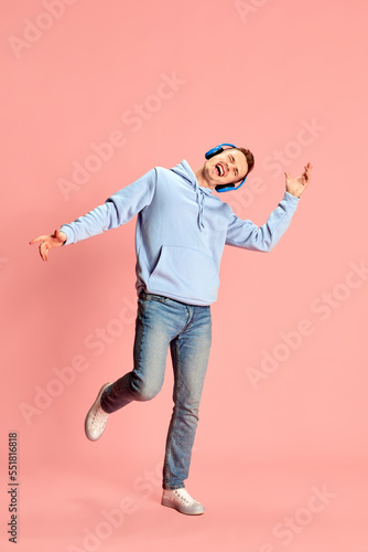 Portrait of young man in hoodie and jeans listening to music in headphones and emotionally singing isolated over pink background. Concept of emotions, music