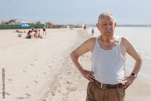 Un signore anziano vestito in canottiera in riva al mare è fermo in piedi con espressione seria