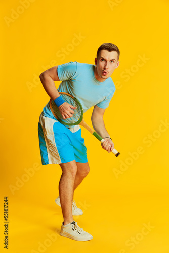 Portrait of young man in blue uniform training, posing with tennis racket isolated over yellow background. Concept of sport and emotions