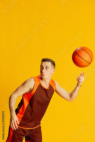 Portrait of young emotive man in orange uniform playing basketball, training, spinning ball on finger isolated over yellow background
