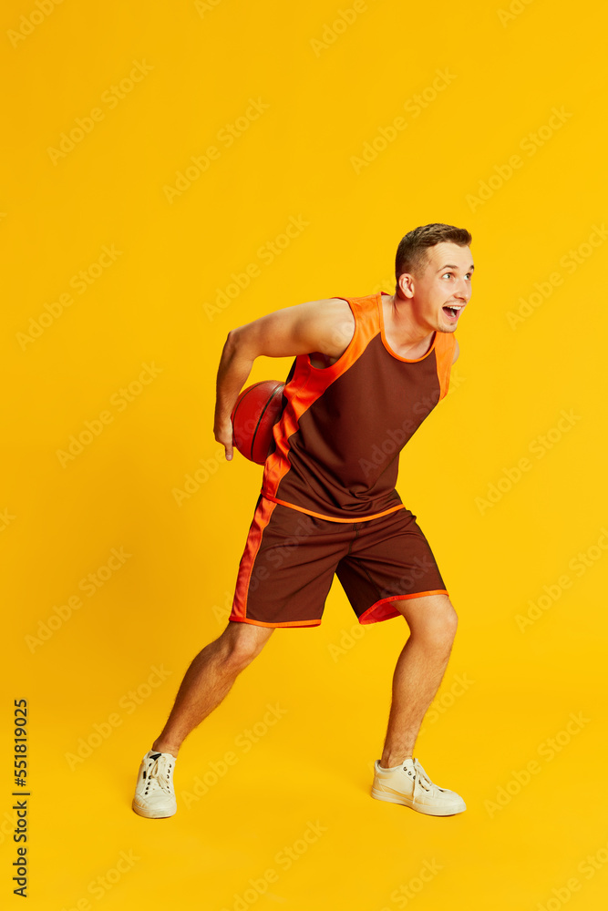 Portrait of young active man in orange uniform training, playing basketball isolated over yellow background. Concept of sport and emotions Portrait of young active man in orange uniform training, playing basketball isolated over yellow background. Concept of sport and emotions