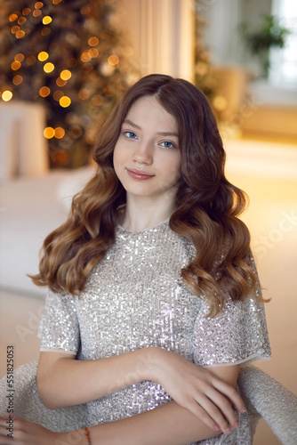 teenage girl is sitting on a chair against the background of a Christmas tree at home in a sequins dress.