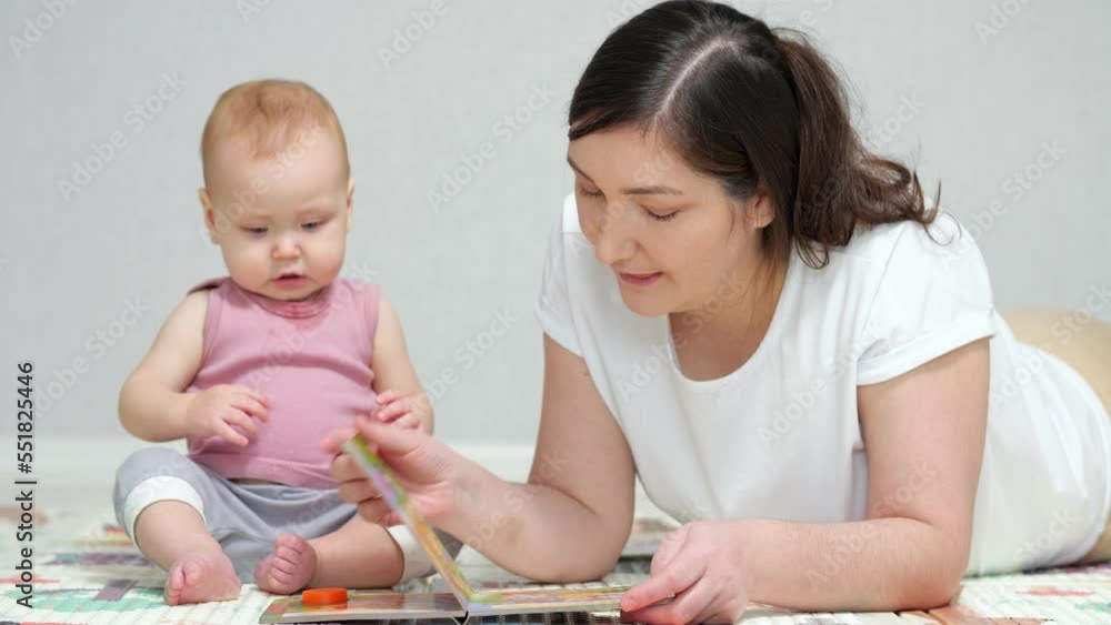 Brown-haired mother reads interesting book to amused baby girl sitting on floor. Child with concerned expression enjoys listening to woman reading, closeup Brown-haired mother reads interesting book to amused baby girl sitting on floor. Child with concerned expression enjoys listening to woman reading, closeup