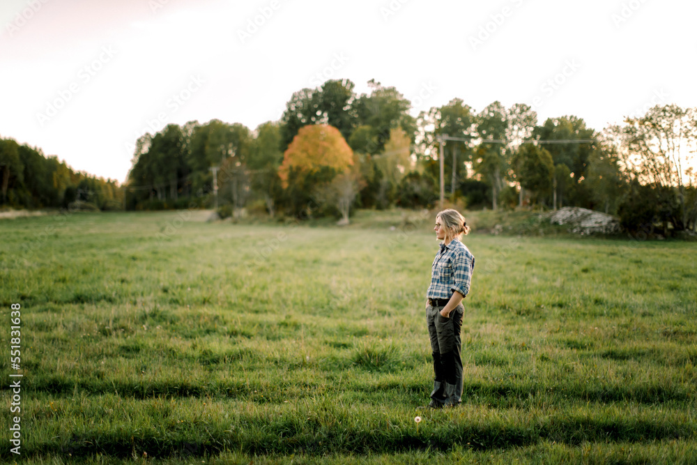 Female farmer with hands in pockets standing on field during sunset Female farmer with hands in pockets standing on field during sunset
