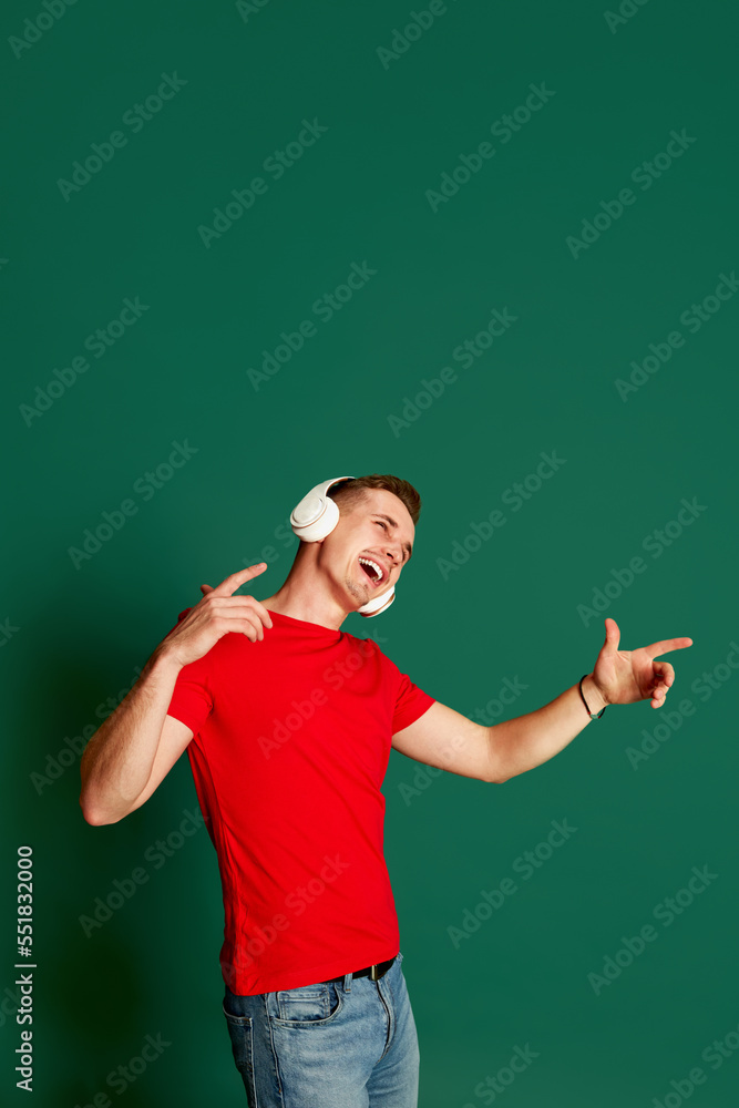 Portrait of young emotive man in casual clothes posing in headphones, singing isolated over green background. Concept of emotions and music Portrait of young emotive man in casual clothes posing in headphones, singing isolated over green background. Concept of emotions and music