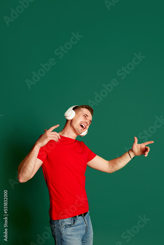 Portrait of young emotive man in casual clothes posing in headphones, singing isolated over green background. Concept of emotions and music