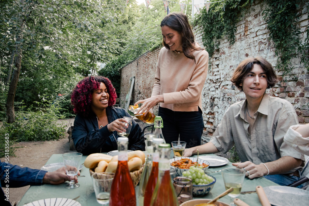 Woman serving drink to female friend sitting at dining table during dinner party in back yard Woman serving drink to female friend sitting at dining table during dinner party in back yard