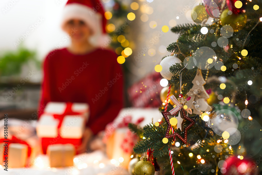 Defocus image of a girl with Christmas presents and a Christmas tree in the foreground. Christmas tree with highlights, blurred lights, gifts and a girl in a red sweater and cap Defocus image of a girl with Christmas presents and a Christmas tree in the foreground. Christmas tree with highlights, blurred lights, gifts and a girl in a red sweater and cap
