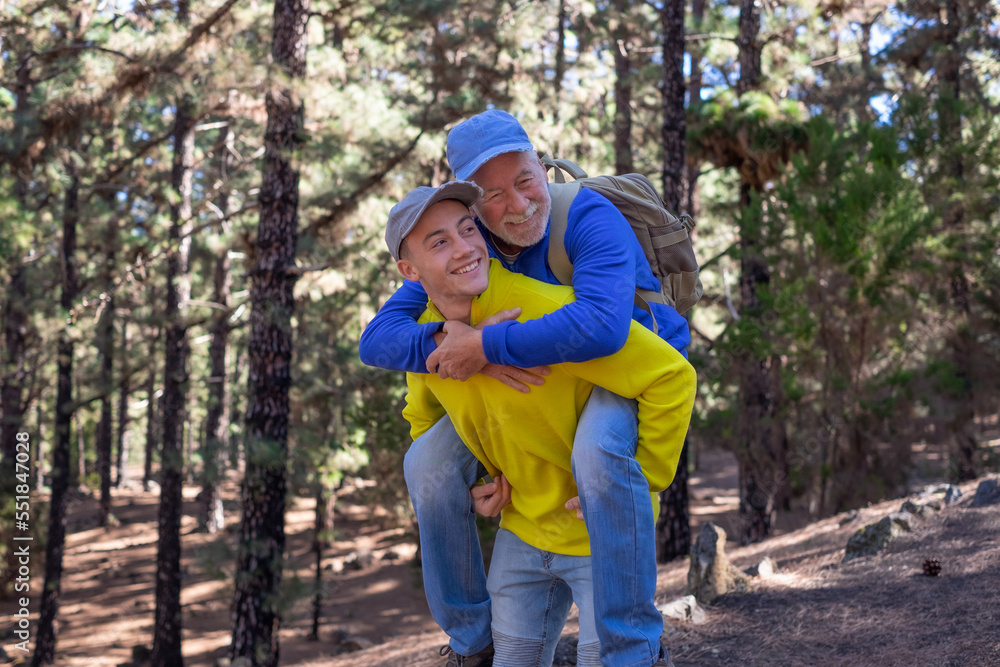 Handsome boy carries his grandfather on shoulders as he walks happily in the forest. Smiling multigeneration family couple enjoying mountain and nature together. The new generation helps the old one Handsome boy carries his grandfather on shoulders as he walks happily in the forest. Smiling multigeneration family couple enjoying mountain and nature together. The new generation helps the old one
