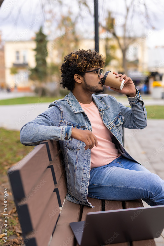 Indian young man sitting on a park bench and drinking coffee while working on laptop in outdoor fresh air at day time. Indian young man sitting on a park bench and drinking coffee while working on laptop in outdoor fresh air at day time.