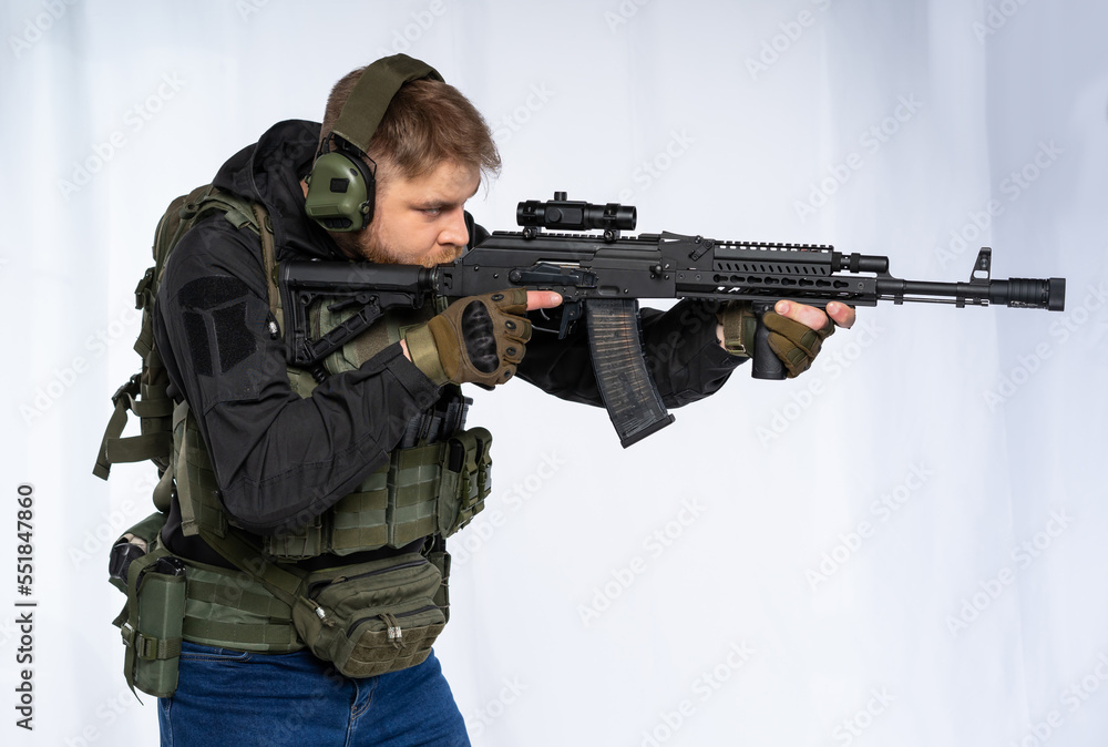 airsoft player in full gear with GG RK74 fire series guns. a man in headphones, a bulletproof vest, with a backpack and a belt, aims his machine gun to the side. profile. White background. airsoft player in full gear with GG RK74 fire series guns. a man in headphones, a bulletproof vest, with a backpack and a belt, aims his machine gun to the side. profile. White background.