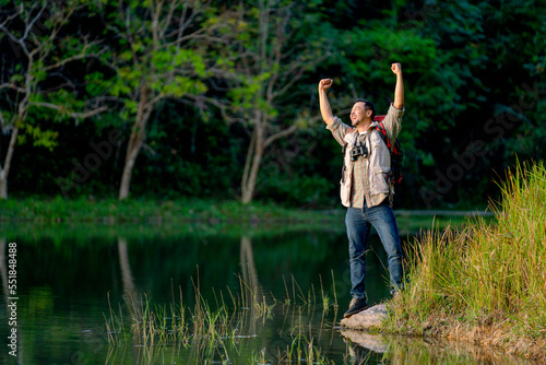 Asian man with bag and journey accessories action of hands up and look happy stand near lake in national park.