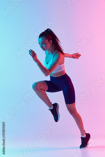 Portrait of young sportive woman training, doing running exercises isolated over gradient blue pink background in neon light. Concept of sport, fitness, health