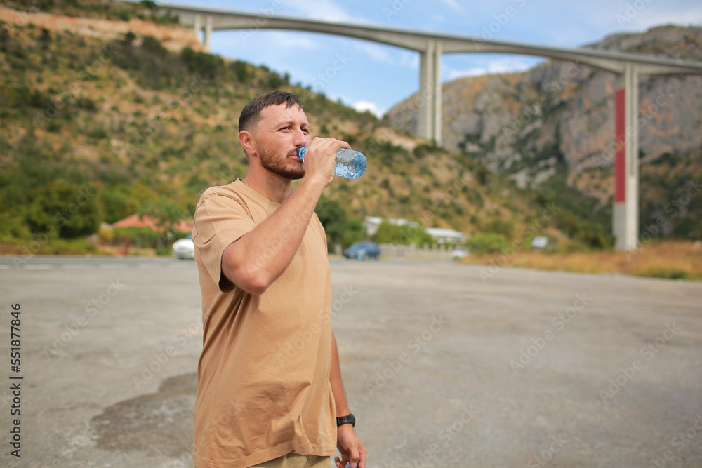 Obraz premium Confident young handsome man with beard drinking water from plastic bottle and enjoying mountains view at hot summer day. Healthcare concept. healthy lifestyle. Refreshing drink