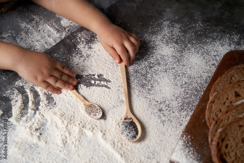 The hands of a kid who helps to make pastries . The child holds wooden spoons with seeds, flour is scattered on the table. High angle. The hands of a kid who helps to make pastries . The child holds wooden spoons with seeds, flour is scattered on the table. High angle.