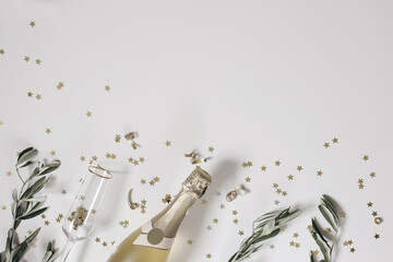 New Year festive web banner. Birthday, wedding party. Celebration concept. Champagne wine bottle, drinking glass. Olive tree branches. White table background. Golden star confetti. Flat lay, top view.