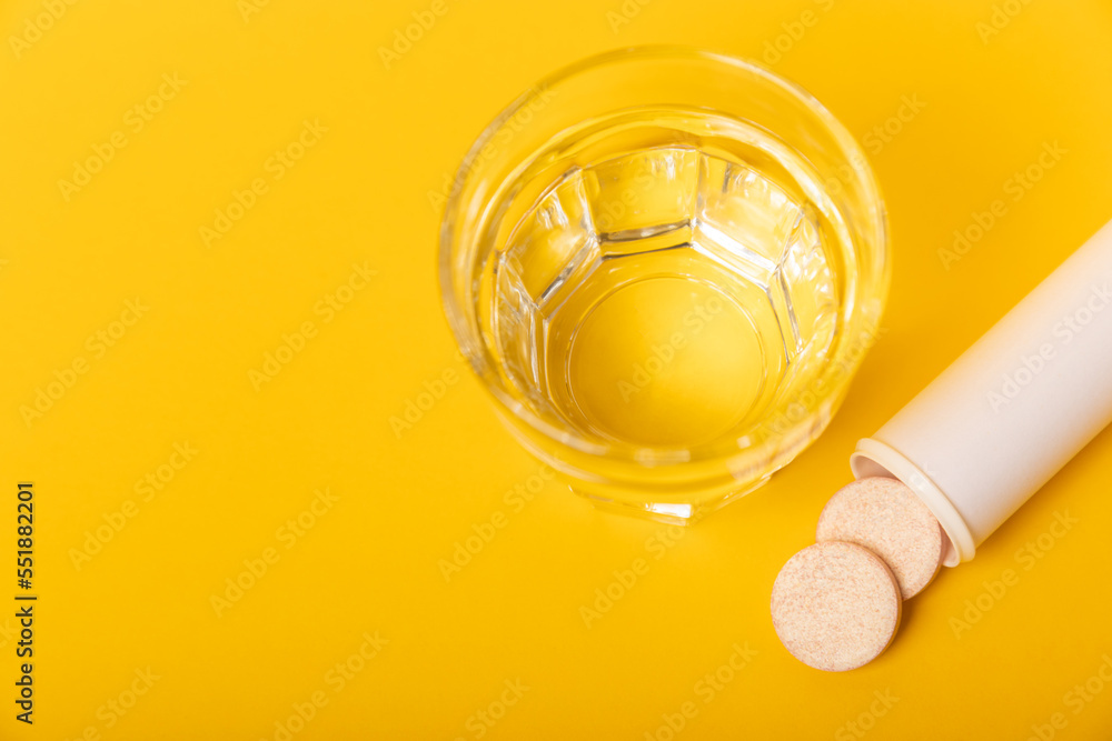 An effervescent vitamin tablet drops and dissolves in a glass of water on a yellow background. The concept of health. Medicine concept. Place for text. An effervescent vitamin tablet drops and dissolves in a glass of water on a yellow background. The concept of health. Medicine concept. Place for text.