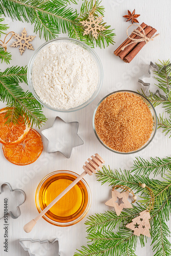 Top view of ingredients for baking of gingerbread biscuit dessert such as honey, flour, sugar and cinnamon arranged on white wooden table with cookie cutters, fir tree branches and christmas decor