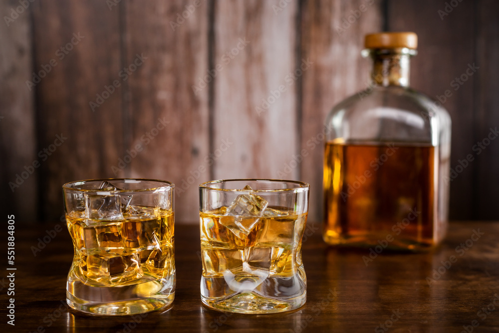 Two glasses of whiskey with ice cubes,with a bottle with whiskey in the background out of focus,on wooden background. Two glasses of whiskey with ice cubes,with a bottle with whiskey in the background out of focus,on wooden background.