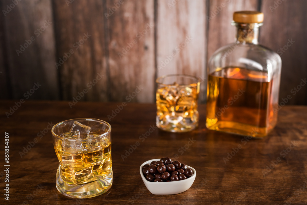 A glass of whiskey with ice cubes next to a chocolate snack, with a bottle of whiskey and another glass in the background out of focus, on wood background. A glass of whiskey with ice cubes next to a chocolate snack, with a bottle of whiskey and another glass in the background out of focus, on wood background.