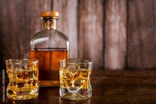 Two glasses of whiskey with ice cubes,with a bottle with whiskey in the background out of focus,on wooden background.