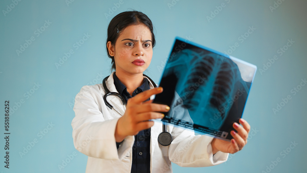 Portrait of a young female doctor examining patient's x-rays scan, Cheerful Asian Indian woman doctor in apron and stethoscope isolated over blue studio background Portrait of a young female doctor examining patient's x-rays scan, Cheerful Asian Indian woman doctor in apron and stethoscope isolated over blue studio background