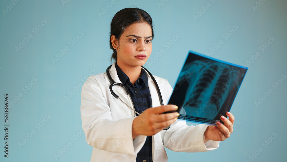 Portrait of a young female doctor examining patient's x-rays scan, Cheerful Asian Indian woman doctor in apron and stethoscope isolated over blue studio background Portrait of a young female doctor examining patient's x-rays scan, Cheerful Asian Indian woman doctor in apron and stethoscope isolated over blue studio background