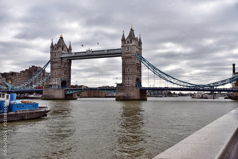 View on the Tower Bridge and the river Thames in London below dark clouds from promenade. There are also moored vessel. Thames river itself looks turbid.