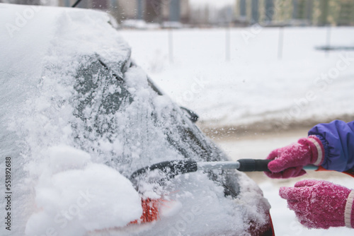 Process of cleaning a car from snow in the morning, girl removing snow from windscreen with a window scraper brush after snowstorm, auto covered in a snow on a parking lot, winter time