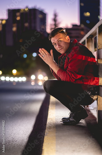 Young and attractive model guy with red jacket on a bridge in the city downtown during a purple sunset