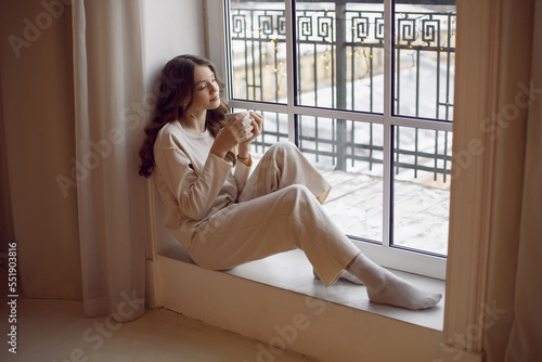 Portrait of a lonely sad teenage girl in a white warm suit sitting on the window with a cup of warm tea in winter at Christmas