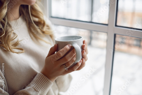 Portrait of a lonely a beautiful blonde woman in a white warm suit sitting on the window with a cup of warm tea in winter at Christmas