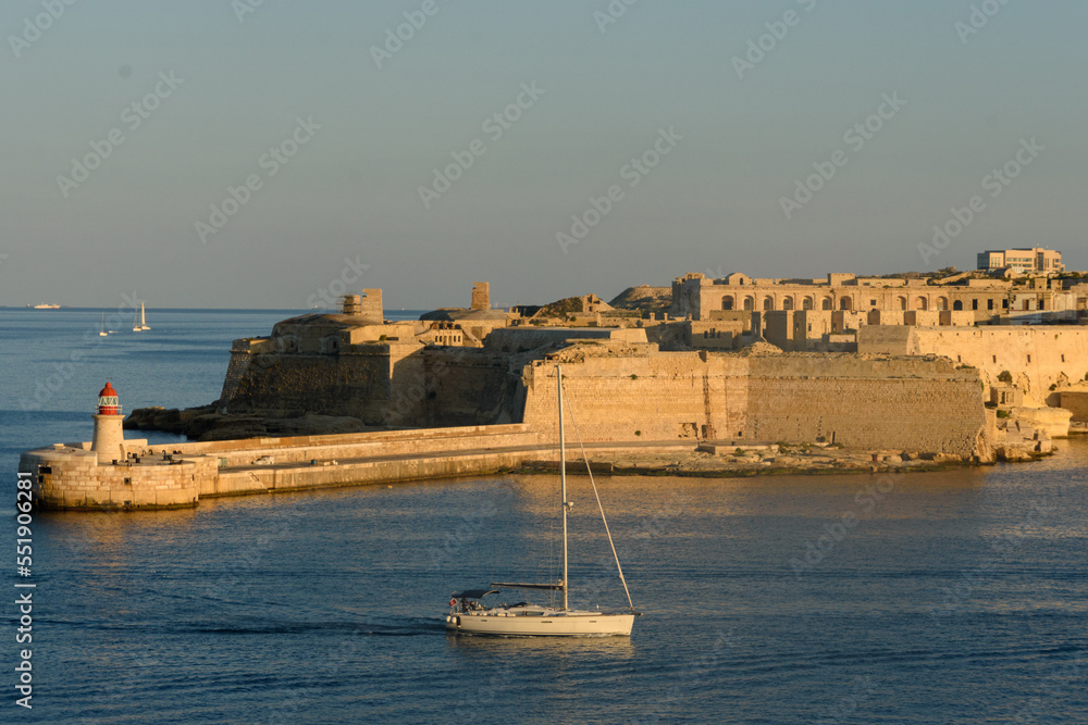A sailing boat entering the Grand Harbour in Malta and passing the lighthouse on the Ricasoli Breakwater and Fort Ricasoli.