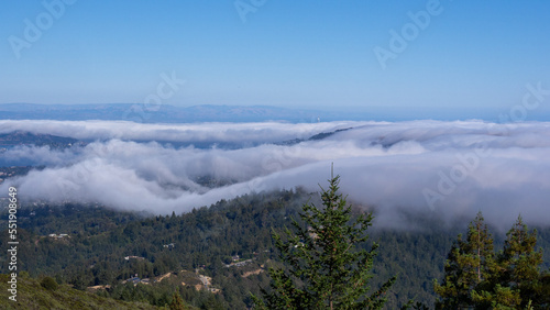 Blue sky and billowing clouds as seen from above with green trees below and a skyscraper in the distance. 