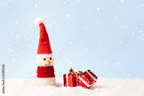 Knitted snowman in a red Christmas hat and sweater with two red gifts. Merry Christmas and New Year 2023 greeting card