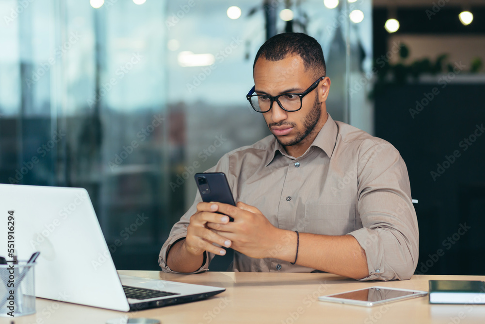 Obraz premium Shocked and surprised young African American man sitting in the office at the table and holding the phone in his hands, received a message, read the news.