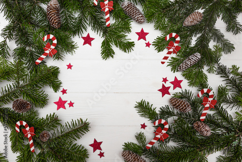 Green Christmas tree branches with pinecones decorated with red stars and Christmas candy canes on a white wooden background top view