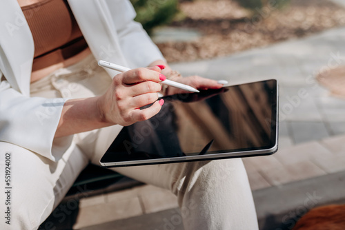 Close up of businesswoman in white suit sitting on electric scooter and working on digital tablet in city with modern architectury photo