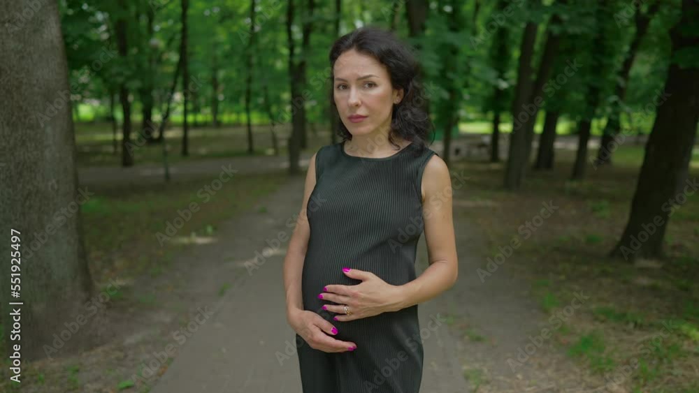 Portrait of depressed pregnant woman in black dress looking at camera with sad facial expression standing on park alley. Frustrated Caucasian lady posing in slow motion on spring summer day outdoors Portrait of depressed pregnant woman in black dress looking at camera with sad facial expression standing on park alley. Frustrated Caucasian lady posing in slow motion on spring summer day outdoors