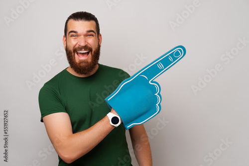 Wide smiling man is wearing blue fan glove and pointing aside with it over grey background. photo